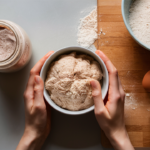 Overhead view of a kitchen counter with a jar of sourdough starter, a bowl of sourdough discard, and baking ingredients.