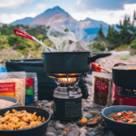 Hiker preparing backpacking meals over a portable stove with a mountain backdrop.